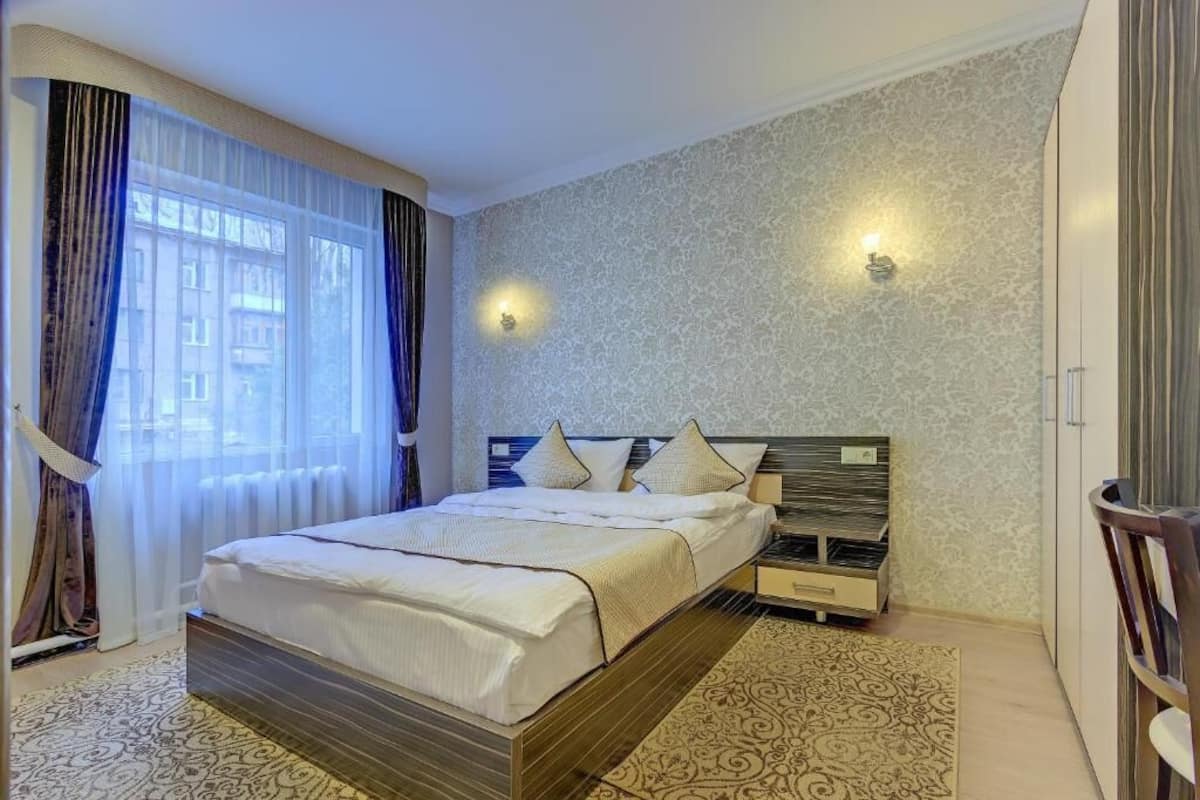 Habitación de hotel con cama, mesita de noche, lámpara y vista a edificios a través de la ventana.