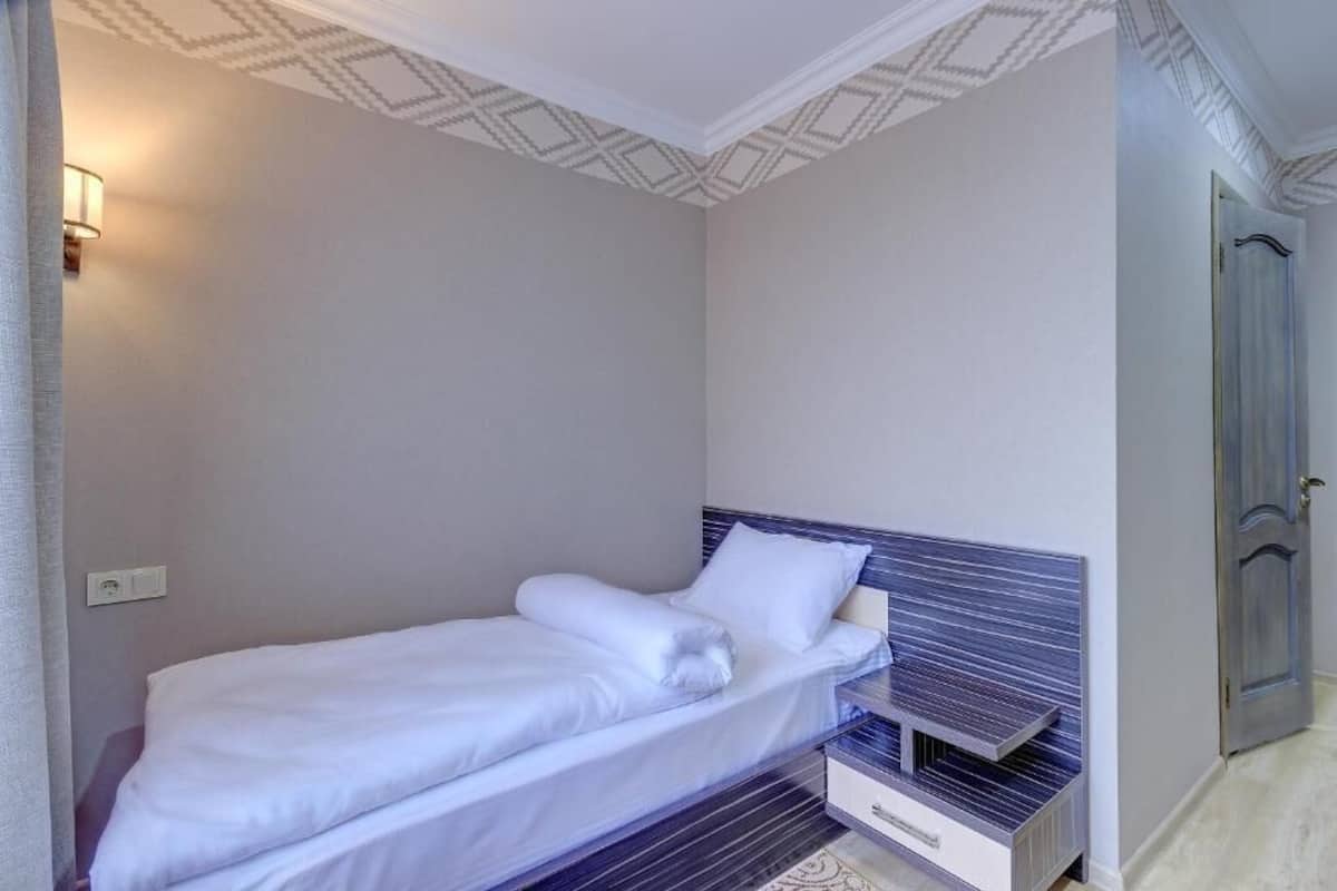 Una cama individual con cabecera, una mesita de noche y un aplique en una habitación con techo estampado y una puerta a la derecha.