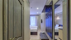 A hotel room with a wooden door, a bed, a bench, a mirror, and a wall-mounted light fixture. - Resident Hotel Almaty (Almaty)