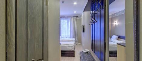A hotel room with a wooden door, a bed, a bench, a mirror, and a wall-mounted light fixture.