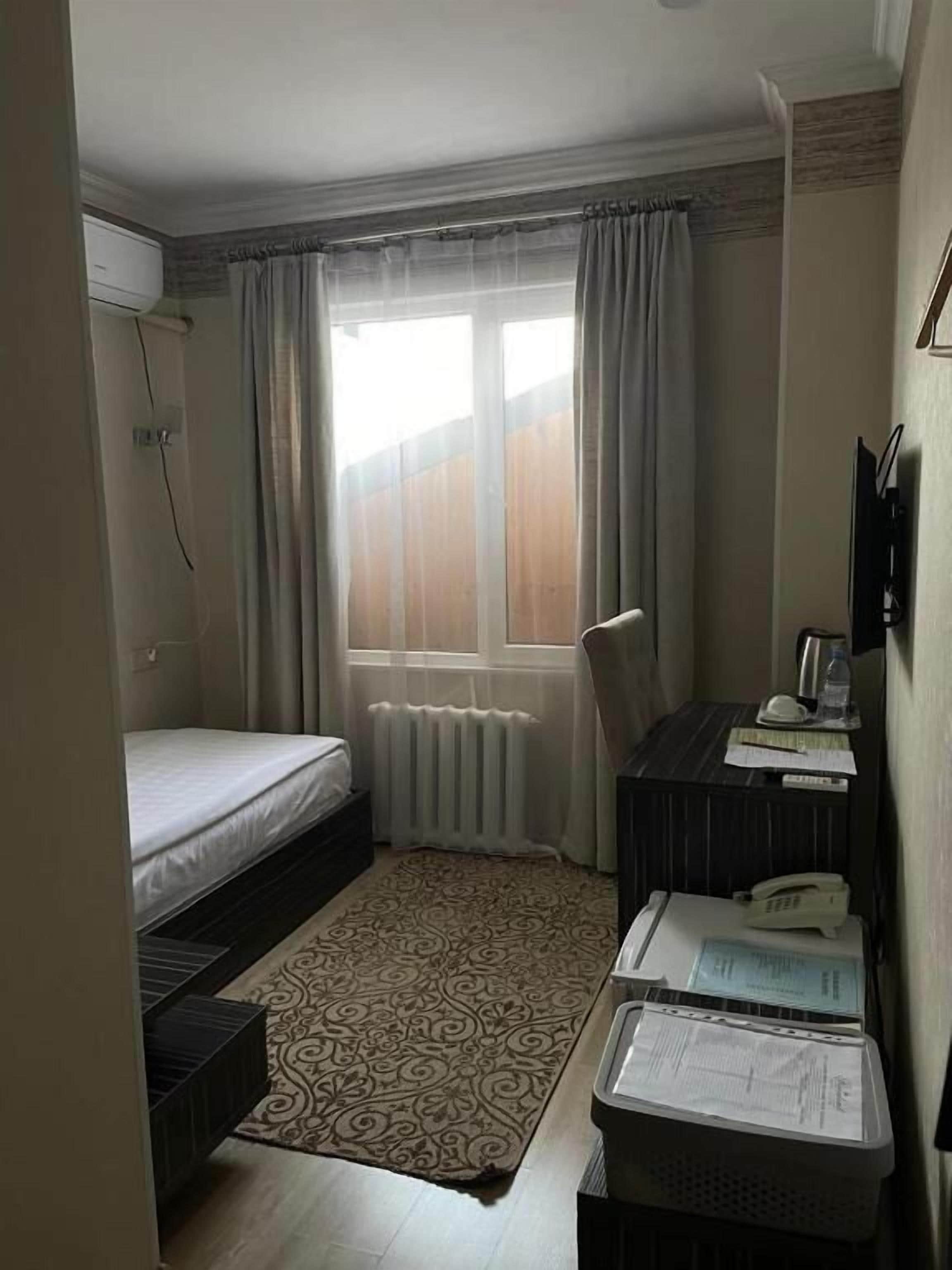 A small, compact room with a bed, a desk, a radiator, and a window with curtains.