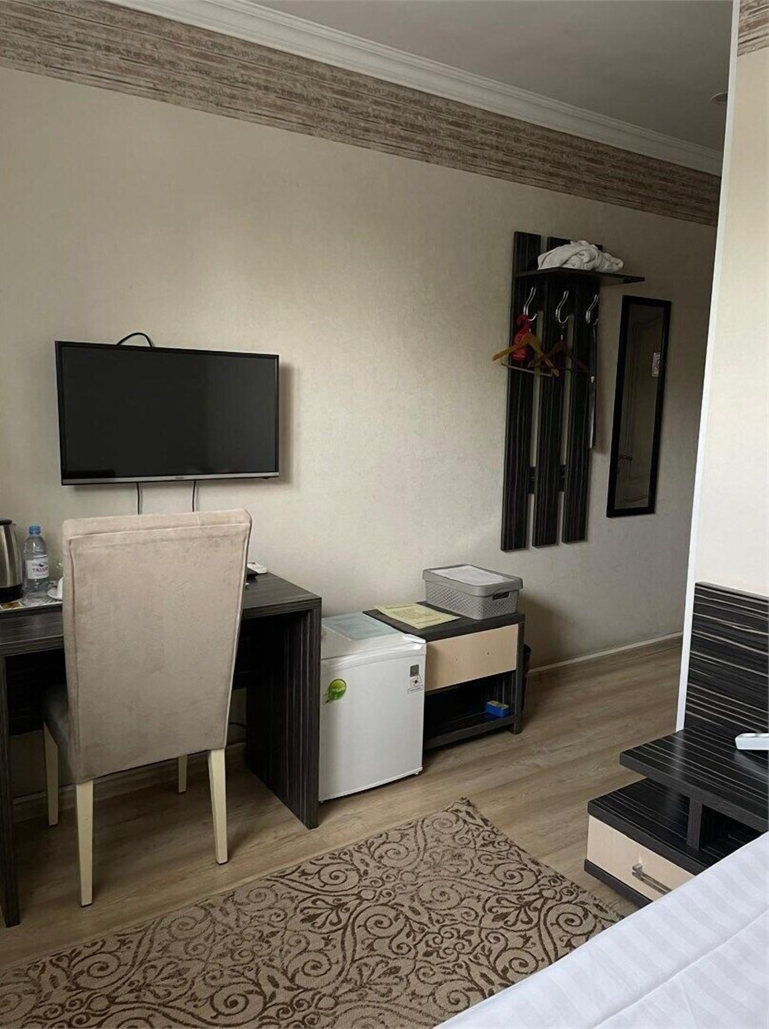 A hotel room with a bed, a desk with a chair, a television, a refrigerator, a small heater, a mirror, and a coat rack with clothes.