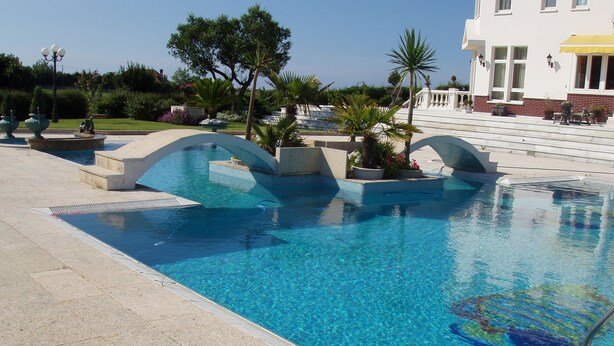 Ein Pool mit einer Brücke, umgeben von einem angelegten Garten und einem Gebäude mit einem gelben Sonnenschirm.