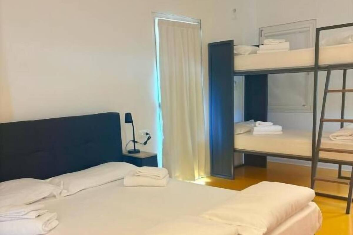 Une chambre d’hôtel avec un lit simple, une table de chevet, une lampe, une fenêtre avec des rideaux et, au fond, un lit superposé.