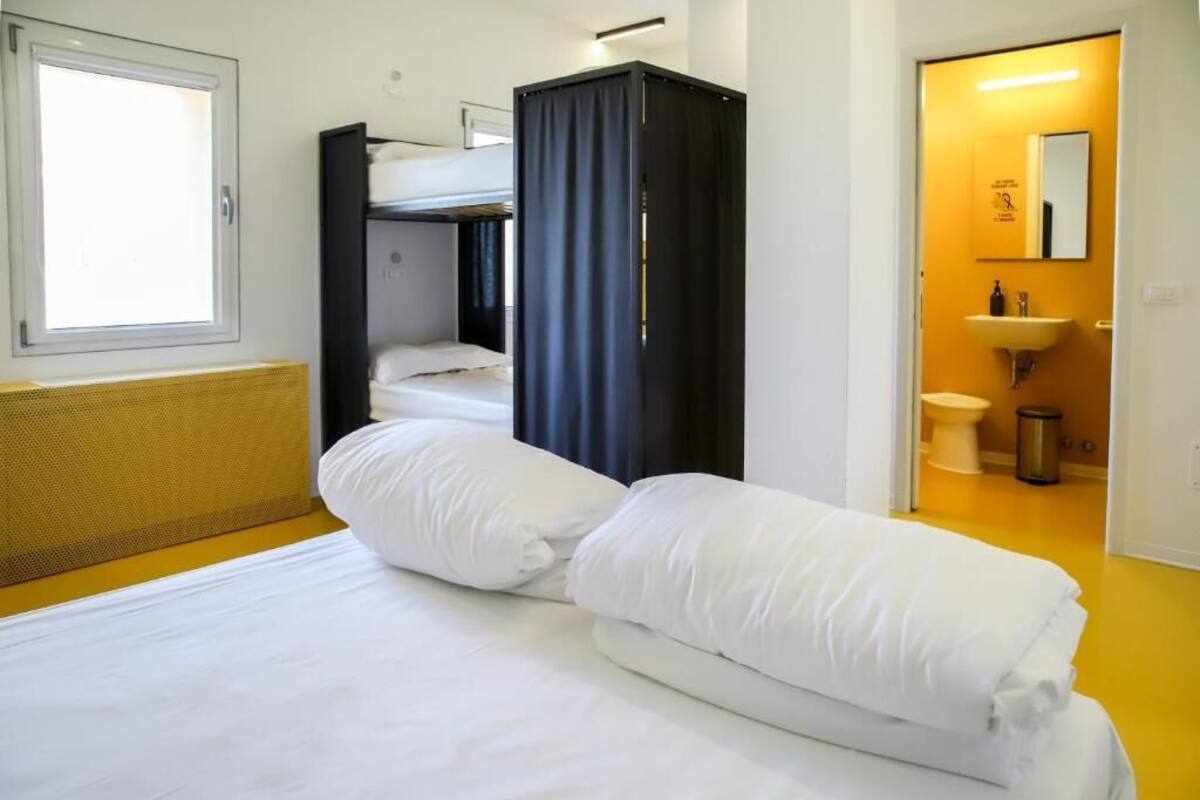 Une chambre d’hôtel avec un lit superposé, un rideau servant de séparation, une petite salle de bain avec un lavabo et des toilettes, et une fenêtre laissant entrer la lumière naturelle.