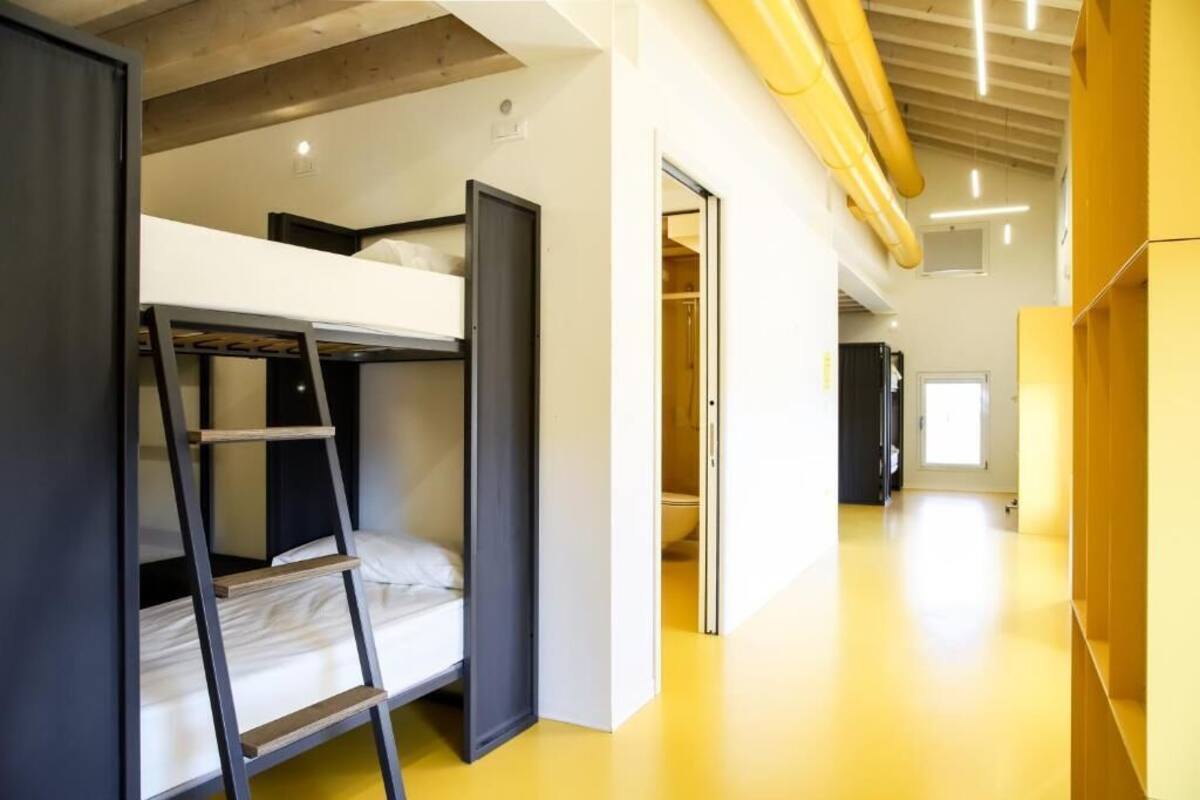 Une chambre avec un lit superposé, une échelle, une porte donnant sur une salle de bain et un couloir aux murs jaunes et aux poutres en bois.