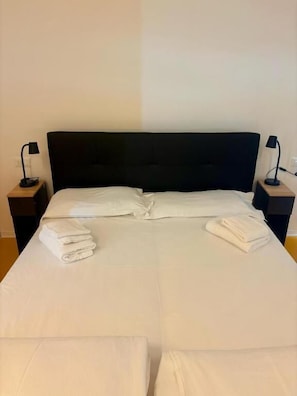 Una cama bien hecha con sábanas blancas, dos mesitas de noche con lámparas y una pared lisa.