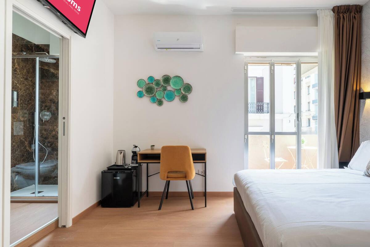 Habitación de hotel con cama, escritorio, silla, refrigerador pequeño, aire acondicionado de pared y un adorno en la pared.