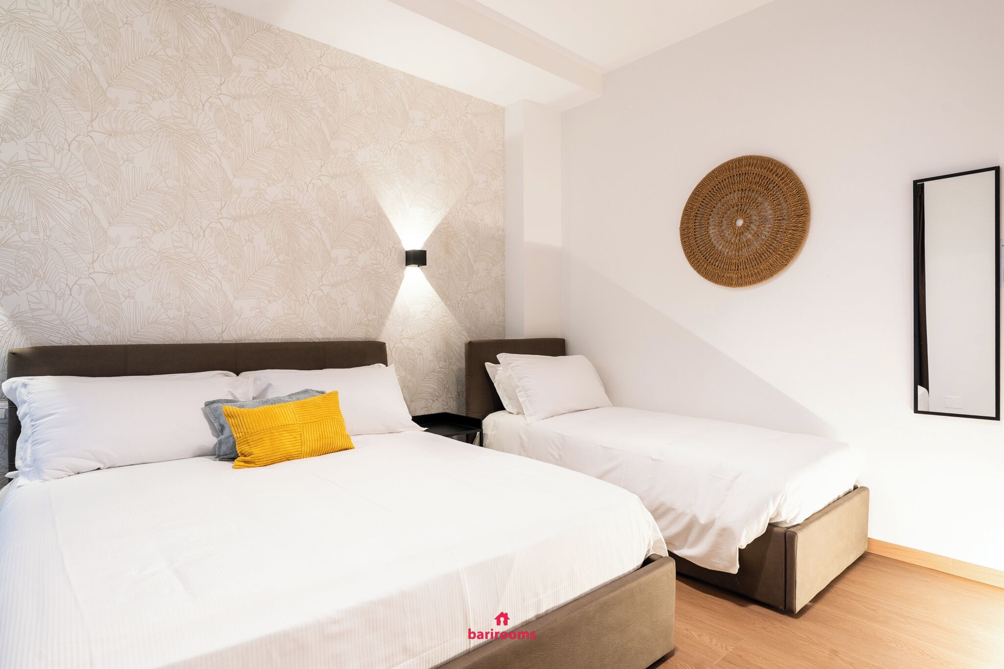 A hotel room with two beds, a textured wall, a wall-mounted light, a woven wall decoration, and a framed picture.