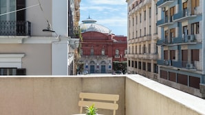 Un balcón con una mesa y una silla blancos, una planta en maceta y vistas de un edificio rojo con una cúpula.