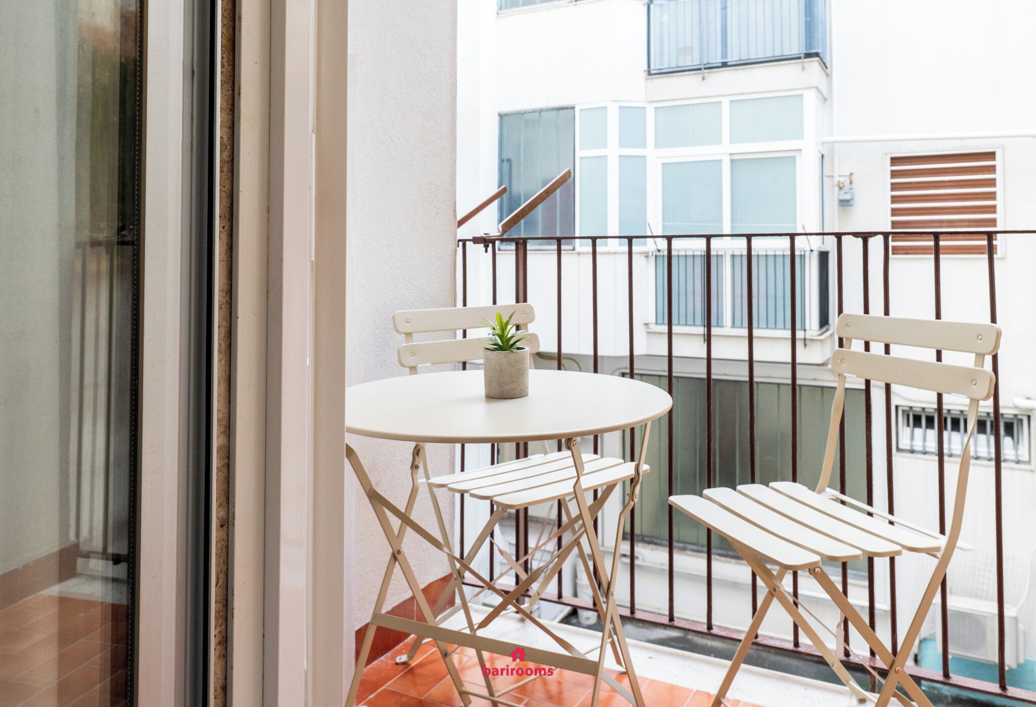 A balcony with a round table and two chairs, a potted plant, and a view of a building with windows and a staircase.