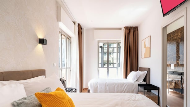 Habitación de hotel con dos camas, televisión, ventana con cortinas y vistas a edificios.