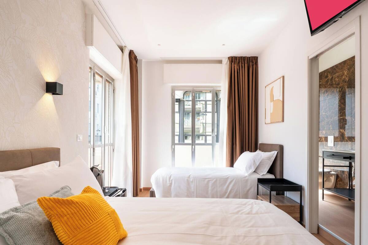 Habitación de hotel con dos camas, televisión, ventana con cortinas y vistas a edificios.
