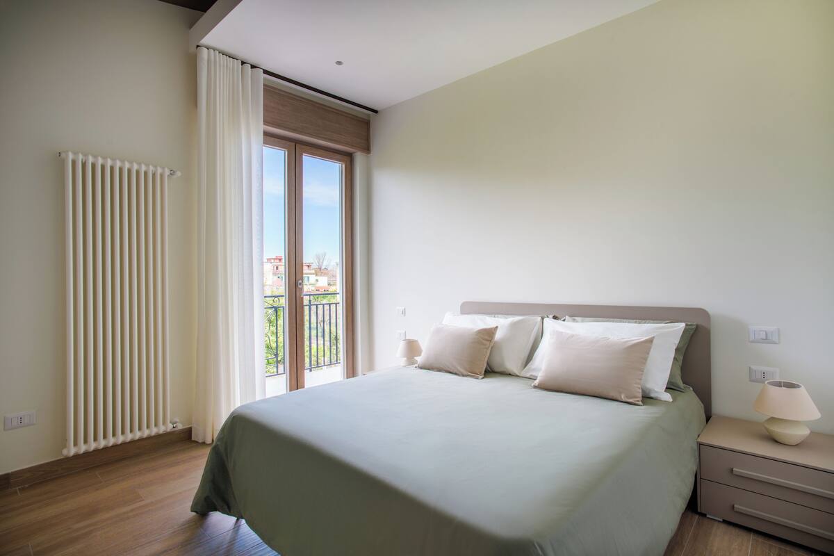 Een slaapkamer met een bed, nachtkastje, lamp, raam met gordijnen en een radiator.