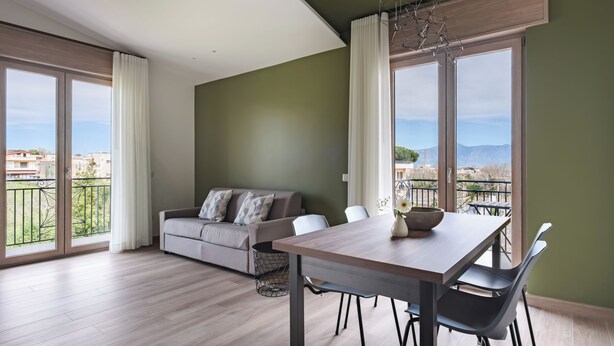 A modern living room with a sofa, dining table, and chairs, large windows with curtains, and a view of the city. - Residenze Vesuviane - Pompei (Terzigno)