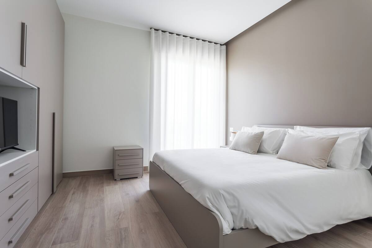 Een slaapkamer met een bed, een televisie, een dressoir en een raam met gordijnen.