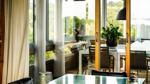 A modern dining area with a glass table, wooden chairs, a potted plant, and large windows offering a view of greenery. - Villa Carla (Bonifacio)