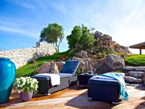 A wooden deck with outdoor seating, a stone wall, and a view of a rocky hillside with trees. - Villa Carla (Bonifacio)