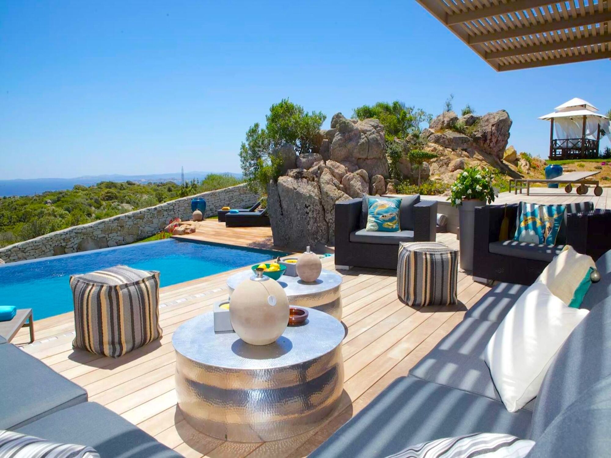 A poolside area with outdoor furniture, a view of the sea, and a rocky landscape.