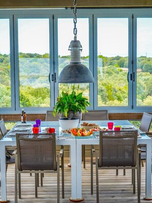 A dining area with a table set for four, a hanging pendant light, and a view of a green landscape through large windows. - Villa Carla (Bonifacio)