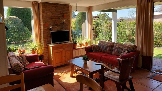 A living room with a TV, sofa, and chairs, a brick fireplace, and large windows with curtains. - Ferienhaus "Datscha" Oostkapelle,  Zeeland,  NL
Familienurlaub (Oostkapelle)