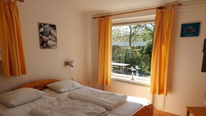 A bedroom with a wooden bed, white bedding, a wall-mounted lamp, a window with curtains, and a small table. - Ferienhaus "Datscha" Oostkapelle,  Zeeland,  NL
Familienurlaub (Oostkapelle)