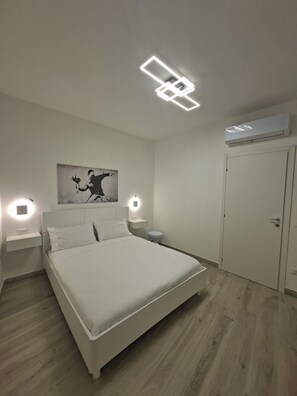 A bedroom with a bed, bedside tables, a wall-mounted light, an air conditioning unit, and a door. - The Little White House: Your elegant suite in the heart of downtown Tuscania (Tuscania)