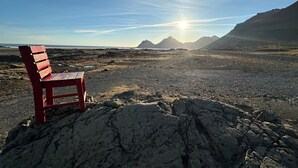 Un banc rouge sur une plage rocheuse, sous un ciel dégagé, avec des montagnes à l’horizon.