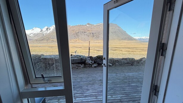 Une vue à travers une porte vitrée donnant sur une terrasse en bois, avec un paysage montagneux en arrière-plan.