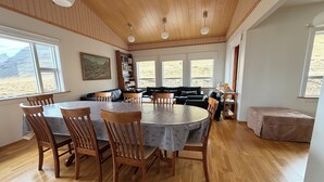 A dining area with a round table and chairs, a black sofa, a bookshelf, and large windows with a view of a mountainous landscape.