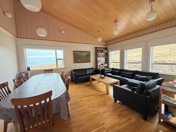A spacious living area with a wooden ceiling, a dining table with chairs, a black leather sofa, a coffee table, and a bookshelf.