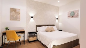 Un dormitorio con cama, mesitas de noche, un escritorio, una silla e iluminación en la pared.