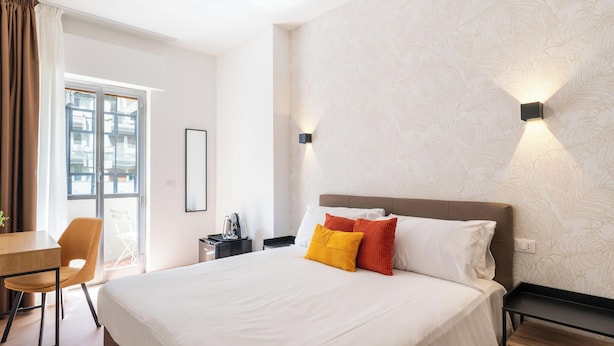 Habitación de hotel con cama, escritorio, silla, mesita de noche y lámpara de pared.