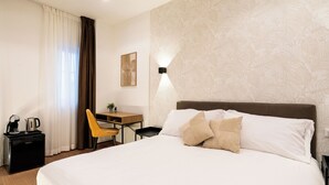 Una habitación de hotel con cama, escritorio, silla, cafetera y una lámpara de pared.