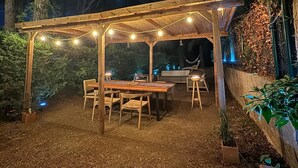 Una veranda esterna con una pergola in legno, luci a catena e un tavolo da pranzo con sedie.