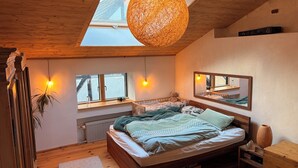 Quarto com teto inclinado, claraboia, piso de madeira, cama com cabeceira, mesas de cabeceira, espelho e janela.