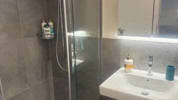 A bathroom with a glass shower enclosure, a white sink with a soap dispenser, and a blue cup on the counter.