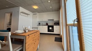 Een kleine keuken met witte kasten, een houten eettafel en een raam met zonwering.