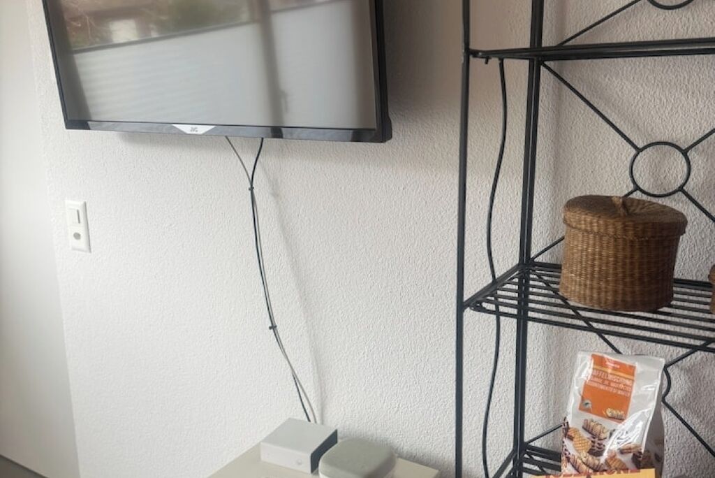 Seinälle asennettu litteä televisio, pistorasia, musta metallihylly, jossa on rottinkikori ja pussi naposteltavaa, sekä valkoinen laatikko lattialla.