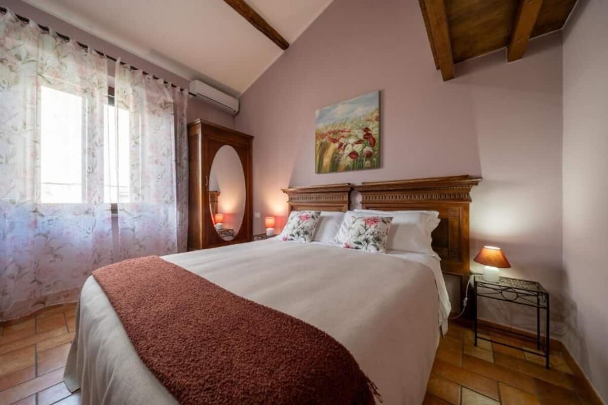 A bedroom with a large bed, a wooden headboard, a painting on the wall, a window with curtains, and a nightstand with a lamp.