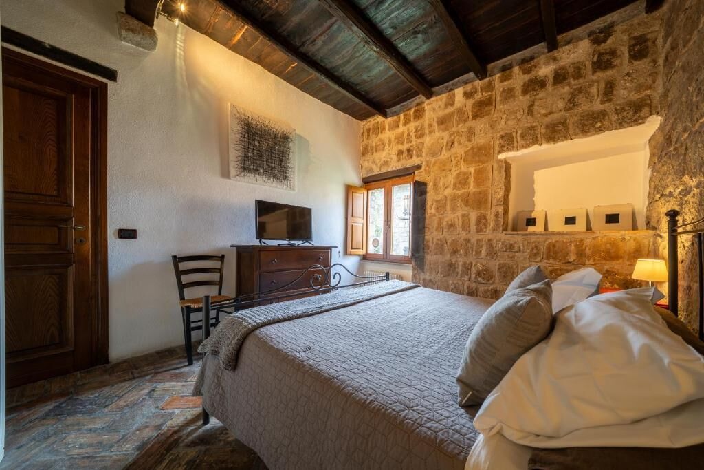 Un dormitorio con pared de piedra, una cama con almohadas, un tocador, un televisor y una ventana con luz natural.