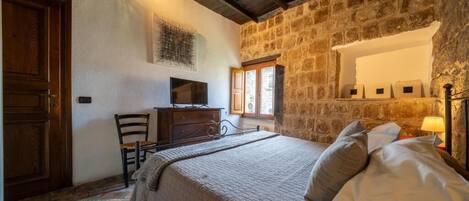 Un dormitorio con pared de piedra, una cama con almohadas, un tocador, un televisor y una ventana con luz natural.