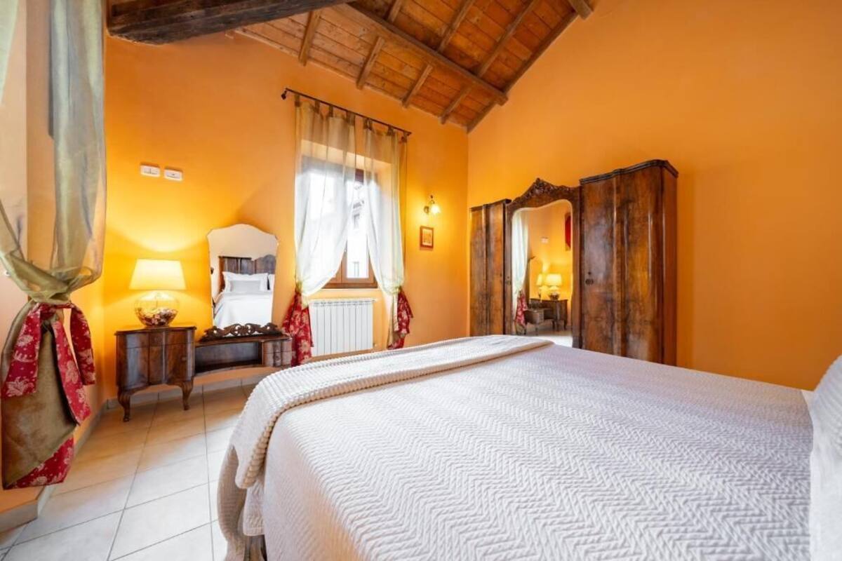 A bedroom with a bed, a wooden wardrobe, a mirror, a nightstand, and a window with curtains.