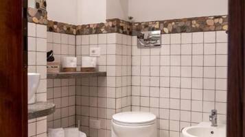 Ein Badezimmer mit Toilette, Bidet und einem kleinen Waschbecken. Eine Wand mit Steinverkleidung und ein gefliester Boden sind vorhanden.