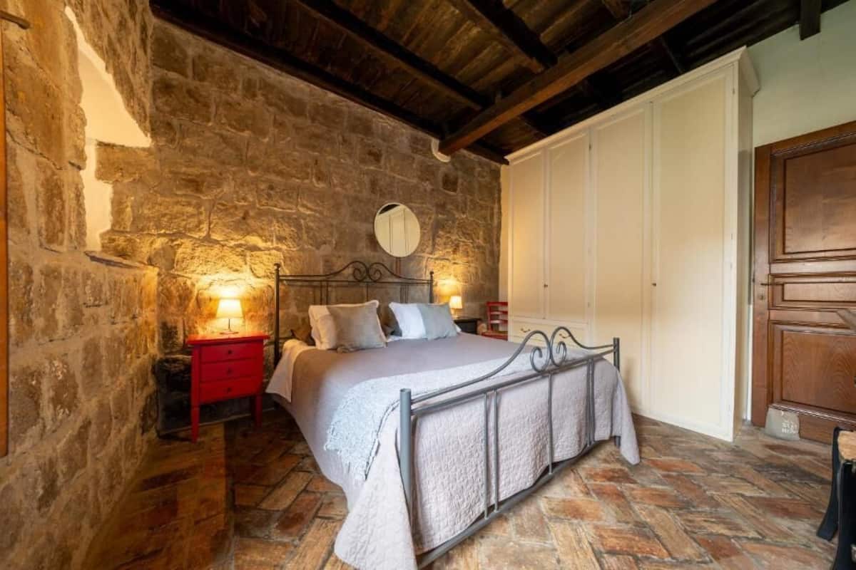 A bedroom with a stone wall, a bed with a metal frame, a red nightstand, and a wooden door.