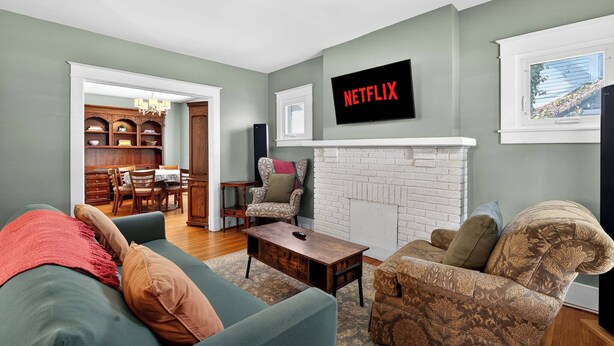 A living room with a fireplace, a television mounted on the wall, and a dining area visible through an open door. - Spacious home w/ yard near Downtown attractions! (Louisville)