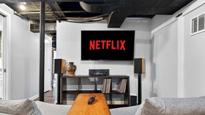 A modern living room with a large flat-screen TV displaying 'NETFLIX,' a wooden coffee table, a sofa, and a bookshelf with various items. - Spacious home w/ yard near Downtown attractions! (Louisville)
