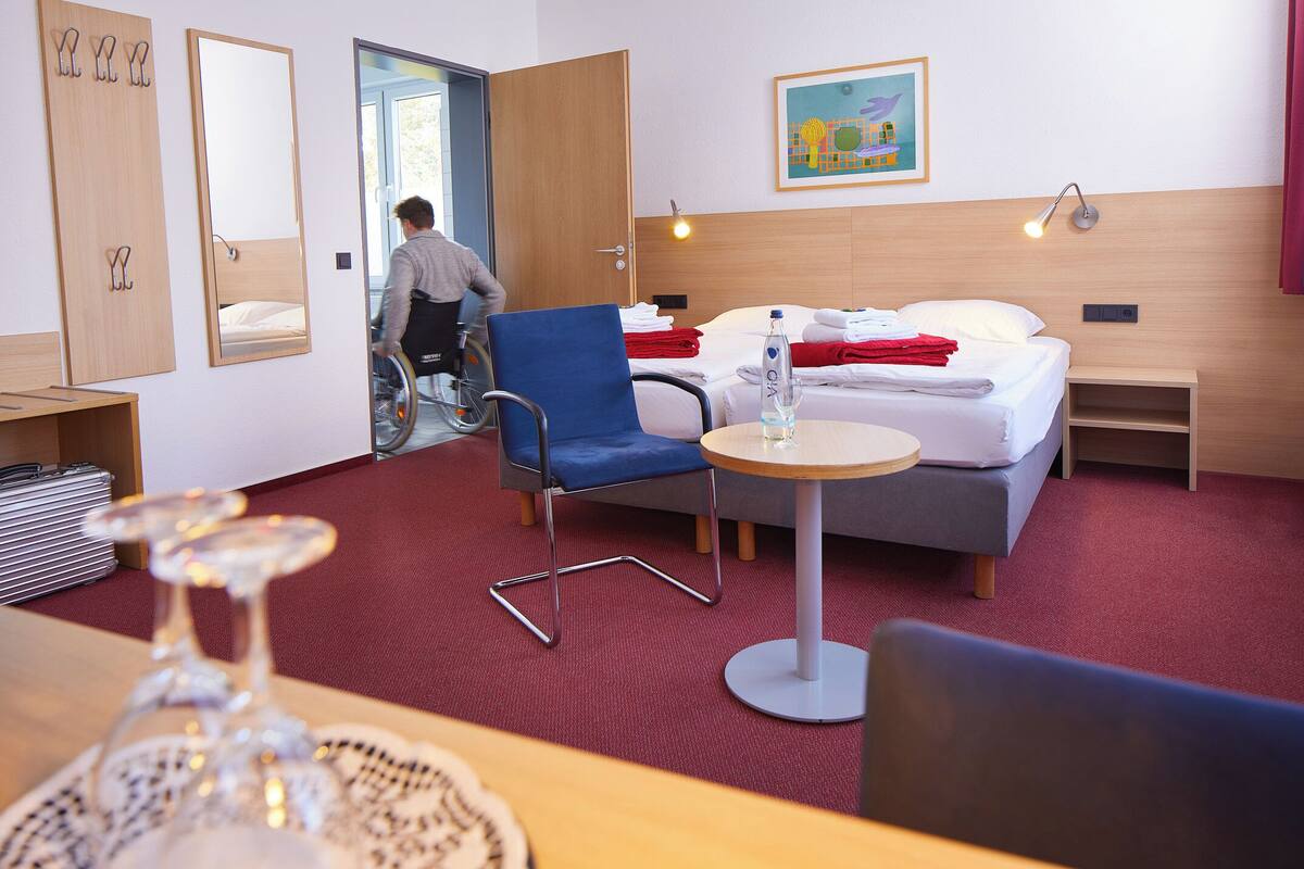 Ein Hotelzimmer mit Rollstuhl, blauem Sessel, Bett mit roten Handtüchern, Nachttisch und Wandlampe.