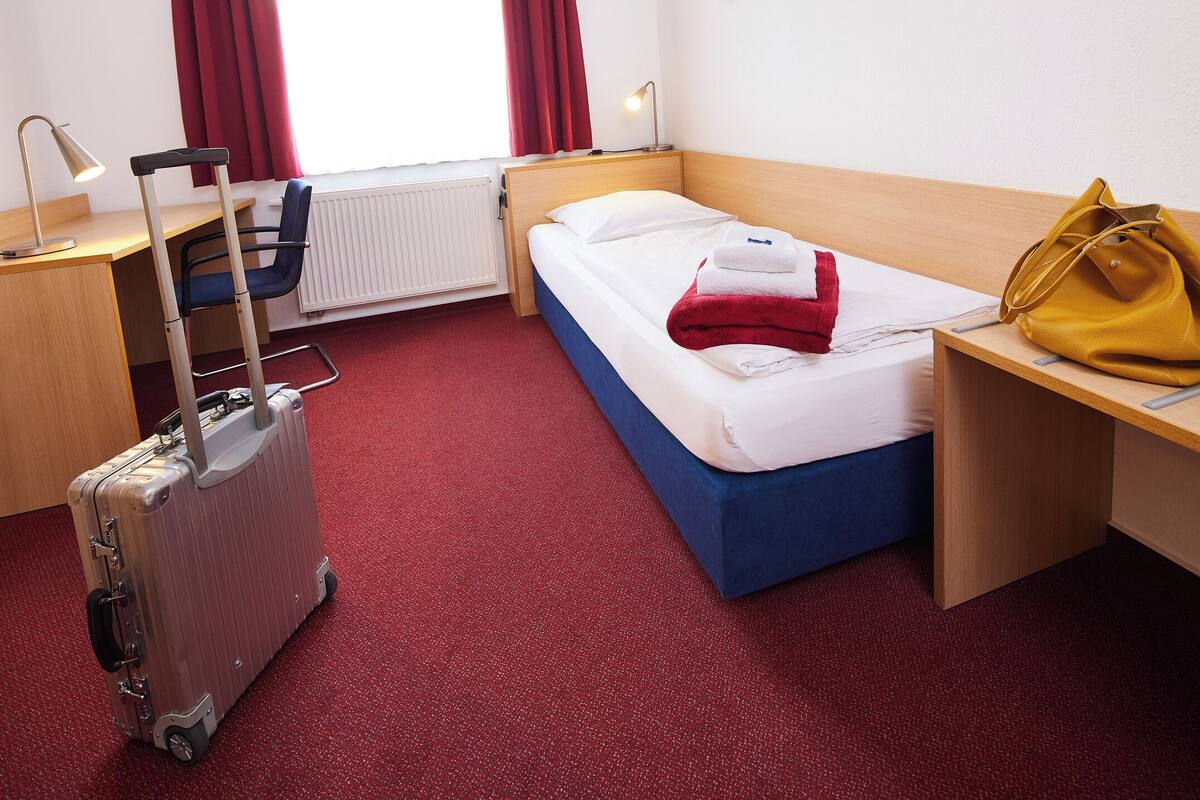 Ein Hotelzimmer mit Bett, Schreibtisch, Stuhl, Koffer und einem Fenster mit Vorhängen.