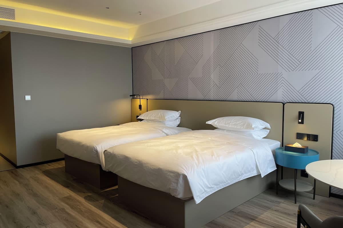 A hotel room with two beds, a bedside table with a lamp, a small round table, and a patterned wall.